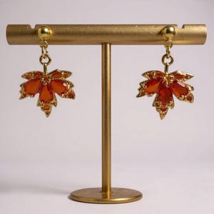 May include: A pair of gold-toned earrings shaped like maple leaves, each adorned with orange-red gemstones. The earrings are displayed on a gold-colored stand, showcasing their intricate design and autumnal color palette. The gemstones are set in a gold frame.