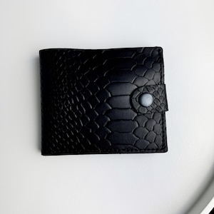 May include: A black bifold wallet featuring a textured snakeskin pattern across the exterior surface. The wallet is secured with a small, circular snap button closure located on the right side, maintaining a sleek and uniform appearance.