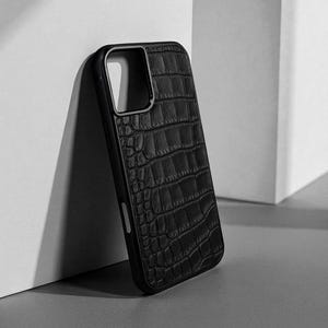 Puede incluir: Una funda para smartphone de color negro con un patrón texturizado de piel de cocodrilo. La funda presenta un recorte rectangular para el objetivo de la cámara en la esquina superior izquierda y un marco negro sólido que rodea los bordes.
