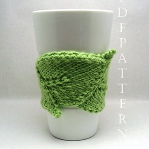 May include: A white ceramic mug with a bright green knitted leaf-shaped cozy wrapped around it. The cozy is made of thick yarn and has a textured appearance. The word "PDFPATTERN" is printed vertically on the right side of the image.