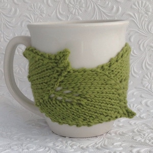 May include: A white ceramic mug with a green knitted cosy. The cosy is shaped like a leaf with a delicate pattern.