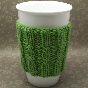 Op de afbeelding: Een witte keramische mok met een groene gebreide cozy. De cozy heeft een kabelbreipatroon.