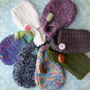 Puede incluir: Una colección de siete bolsas de punto en varios colores y patrones, incluyendo morado, gris, verde, marrón, blanco y rosa. Cada bolsa tiene un diseño único y presenta un botón o un amuleto decorativo.