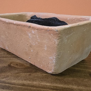 May include: A rectangular, terracotta-colored planter with a dark, charcoal-like object inside. The planter has a rough, textured surface and sits on a dark wooden surface. The background is a solid orange color.