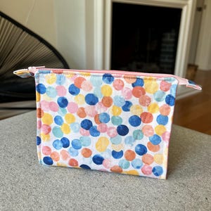 May include: A white zippered pouch with a colorful confetti pattern. The pouch is made of fabric and has a pink zipper.