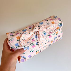 May include: A rolled-up, floral-patterned changing pad in shades of pink, blue, orange, and purple. The pad is tied with a matching bow. The changing pad is designed for babies.