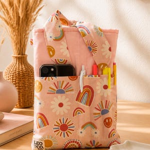 May include: A pink fabric pouch featuring a repeating pattern of rainbows, daisies, and smiley faces. The pouch has a top handle, a front pocket containing a smartphone and several pens, and a small white label reading LEO CO. at the bottom corner.