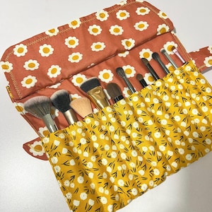 May include: A floral makeup brush roll in rust and yellow. The roll is open, revealing various makeup brushes. The rust-colored flap features a white daisy pattern, while the brush holder is yellow with a white and black floral design.