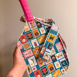 Puede incluir: Una raqueta de tenis con un mango rosa se coloca dentro de una bolsa de tenis de tela. La bolsa presenta un fondo verde azulado con un patrón repetido de diseños de sellos postales coloridos. La bolsa tiene una correa a juego.