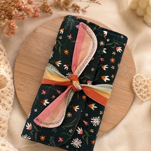 May include: A dark green floral print case tied with a multi-colored ribbon. The case features a repeating pattern of white and orange flowers and green leaves. The ribbon is tied in a bow and has pink, orange, and yellow stripes. The case is on a wooden surface.