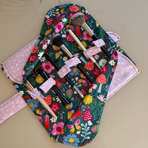 May include: A fabric makeup brush roll with a dark green background featuring a colorful pattern of red mushrooms, yellow butterflies, and various flowers. The interior has light pink polka-dot fabric flaps and five individual slots with light pink snap-button straps to secure five different makeup brushes.