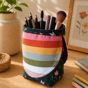 Peut inclure: Une trousse en tissu avec un extérieur à motif floral vert foncé et un panneau avant présentant des rayures horizontales roses, orange, jaunes, bleu clair, vert olive et blanches. La trousse contient divers pinceaux et crayons.