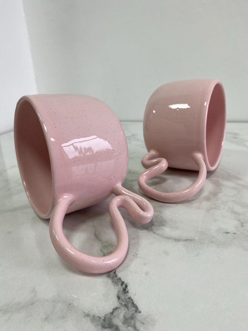 K&ouml;nnte beinhalten: Zwei rosa Keramikbecher mit einzigartigen, geschwungenen Griffen. Die Becher haben eine gesprenkelte Oberfl&auml;che und eine gl&auml;nzende Glasur. Die Griffe haben ein unverwechselbares, geschwungenes Design. Die Becher stehen auf einer marmorierten Oberfl&auml;che.
