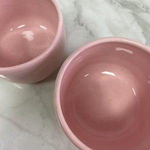 K&ouml;nnte beinhalten: Zwei rosa Keramikschalen mit gesprenkelten Details. Die Schalen haben eine abgerundete Form und eine glatte, gl&auml;nzende Oberfl&auml;che. Die Schalen stehen auf einer wei&szlig;-grauen, marmor&auml;hnlichen Oberfl&auml;che.