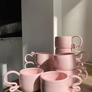 K&ouml;nnte beinhalten: Eine Sammlung rosa Keramikbecher mit einzigartigen, geschwungenen Griffen. Die Becher sind auf einer Holzoberfl&auml;che gestapelt, wobei das Sonnenlicht Schatten wirft. Das Design ist minimalistisch und modern, passend f&uuml;r K&uuml;chenartikel.