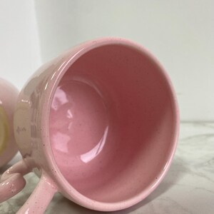 K&ouml;nnte beinhalten: Nahaufnahme einer rosa Keramiktasse mit Henkel. Die Tasse hat eine gesprenkelte Textur und ein gl&auml;nzendes Finish. Das Innere der Tasse ist einfarbig rosa. Der Griff ist gebogen und an der Seite der Tasse befestigt.