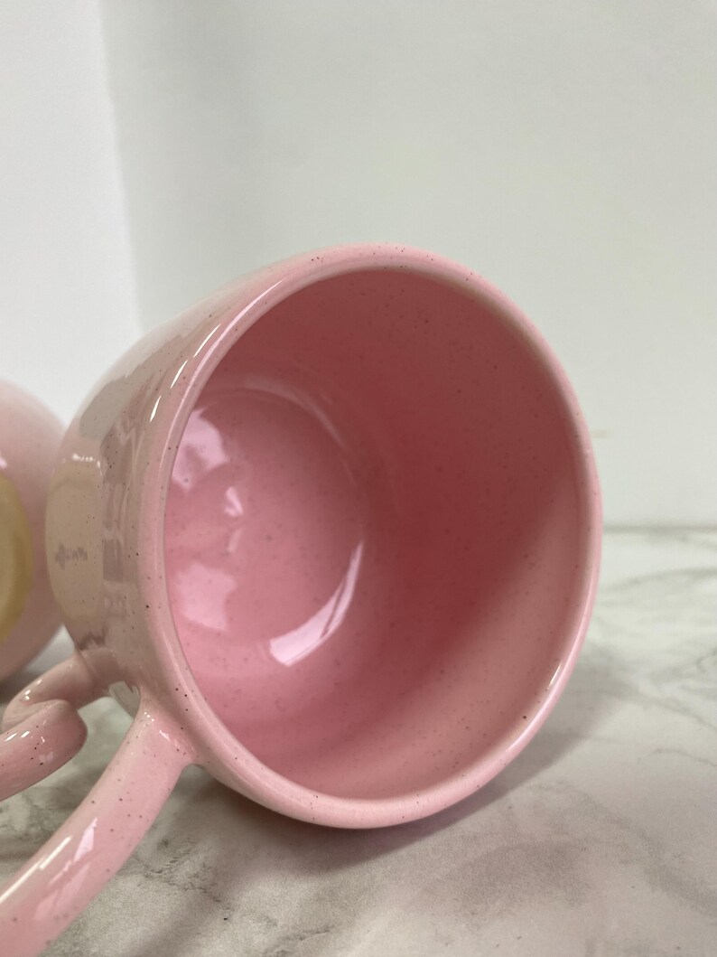 K&ouml;nnte beinhalten: Nahaufnahme einer rosa Keramiktasse mit Henkel. Die Tasse hat eine gesprenkelte Textur und ein gl&auml;nzendes Finish. Das Innere der Tasse ist einfarbig rosa. Der Griff ist gebogen und an der Seite der Tasse befestigt.