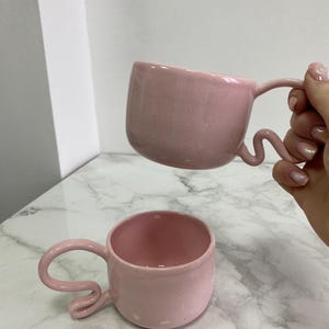 K&ouml;nnte beinhalten: Zwei rosa Keramikbecher mit einzigartigen, wellenf&ouml;rmigen Griffen. Die Becher haben eine gesprenkelte Textur und eine abgerundete Form. Ein Becher wird hochgehalten, w&auml;hrend der andere auf einer marmor&auml;hnlichen Oberfl&auml;che steht. Die Becher sind in einem sanften Pastellrosa gehalten.
