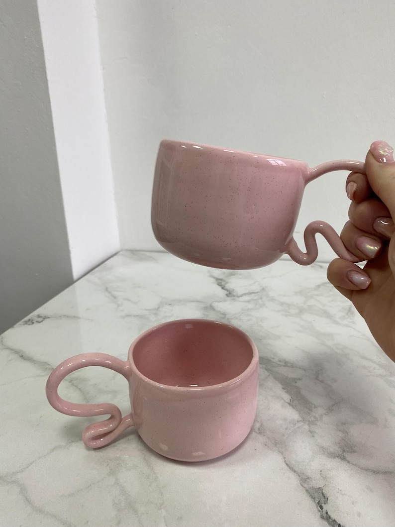 K&ouml;nnte beinhalten: Zwei rosa Keramikbecher mit einzigartigen, wellenf&ouml;rmigen Griffen. Die Becher haben eine gesprenkelte Textur und eine abgerundete Form. Ein Becher wird hochgehalten, w&auml;hrend der andere auf einer marmor&auml;hnlichen Oberfl&auml;che steht. Die Becher sind in einem sanften Pastellrosa gehalten.