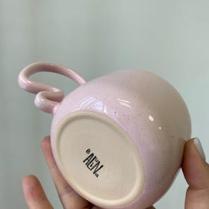 K&ouml;nnte beinhalten: Eine rosa Keramiktasse mit einem einzigartigen Griffdesign. Die Tasse hat eine gesprenkelte Oberfl&auml;che und eine abgerundete Form. Auf dem Boden der Tasse steht der Text "AEN" in Schwarz. Die Tasse wird in einer Hand gehalten.