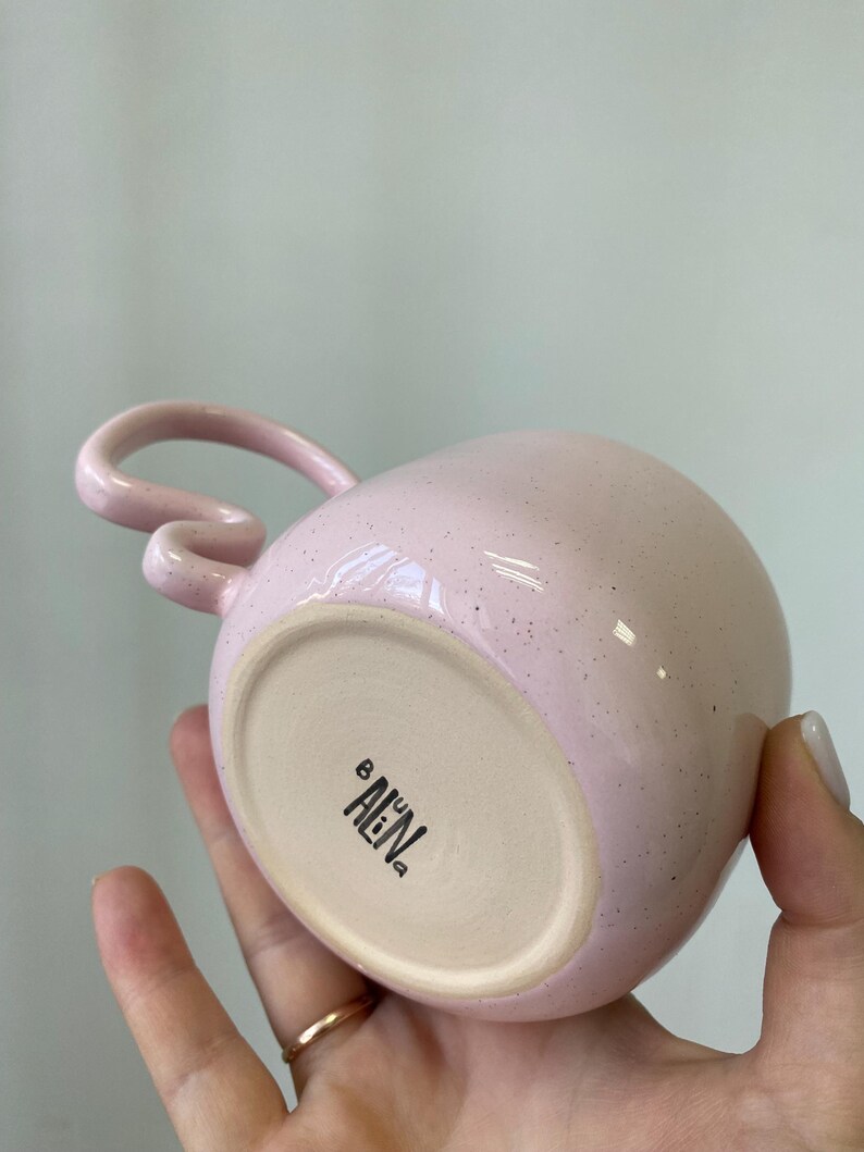 K&ouml;nnte beinhalten: Eine rosa Keramiktasse mit einem einzigartigen Griffdesign. Die Tasse hat eine gesprenkelte Oberfl&auml;che und eine abgerundete Form. Auf dem Boden der Tasse steht der Text "AEN" in Schwarz. Die Tasse wird in einer Hand gehalten.