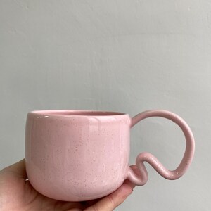 K&ouml;nnte beinhalten: Eine rosa Keramiktasse mit einem einzigartigen, wellenf&ouml;rmigen Griff. Die Tasse hat eine gesprenkelte Textur und einen abgerundeten K&ouml;rper. Der Griff hat ein verspieltes, abstraktes Design. Die Tasse wird in einer Hand vor einem neutralen Hintergrund gehalten.