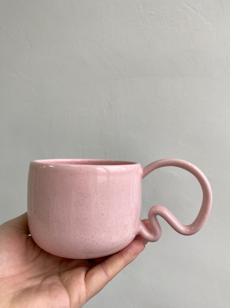K&ouml;nnte beinhalten: Eine rosa Keramiktasse mit einem einzigartigen, wellenf&ouml;rmigen Griff. Die Tasse hat eine gesprenkelte Textur und einen abgerundeten K&ouml;rper. Der Griff hat ein verspieltes, abstraktes Design. Die Tasse wird in einer Hand vor einem neutralen Hintergrund gehalten.