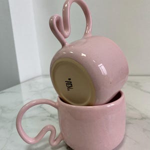 K&ouml;nnte beinhalten: Zwei rosa Keramikbecher, die &uuml;bereinander gestapelt sind und einzigartige, geschwungene Griffe aufweisen. Die Becher haben eine gesprenkelte Textur und ein gl&auml;nzendes Finish. Die Griffe haben ein unverwechselbares, wellenf&ouml;rmiges Design. Der Boden des oberen Bechers ist sichtbar.
