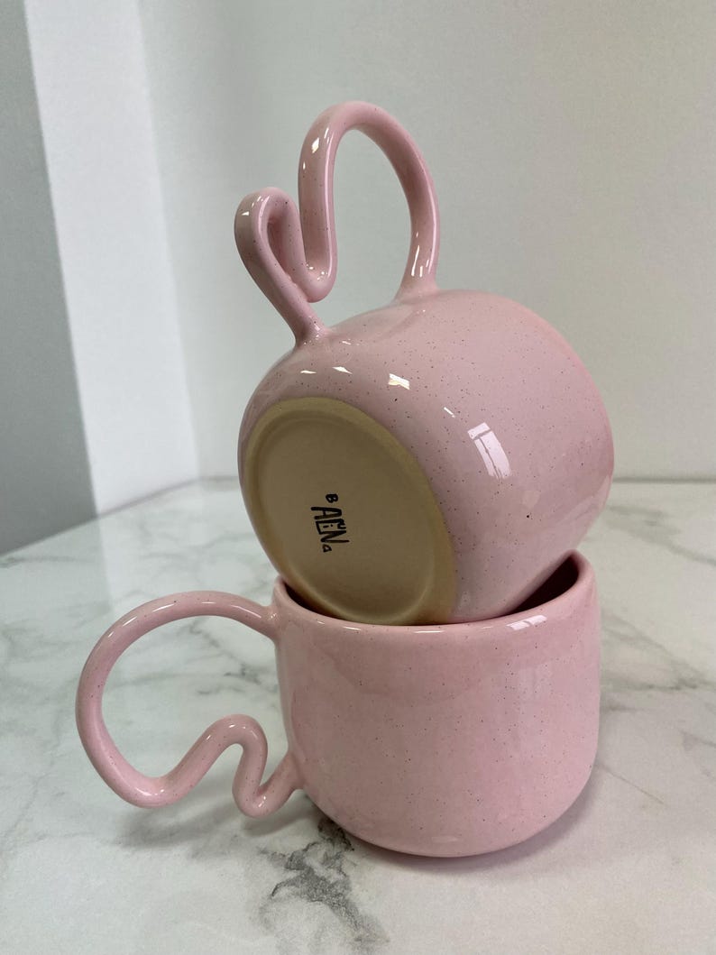 K&ouml;nnte beinhalten: Zwei rosa Keramikbecher, die &uuml;bereinander gestapelt sind und einzigartige, geschwungene Griffe aufweisen. Die Becher haben eine gesprenkelte Textur und ein gl&auml;nzendes Finish. Die Griffe haben ein unverwechselbares, wellenf&ouml;rmiges Design. Der Boden des oberen Bechers ist sichtbar.