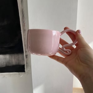 K&ouml;nnte beinhalten: Eine rosa Keramiktasse mit quadratischem Korpus und einem einzigartigen, gewirbelten Henkel. Die Tasse hat eine gesprenkelte Textur und ein gl&auml;nzendes Finish. Die Tasse wird in einer Hand gehalten.