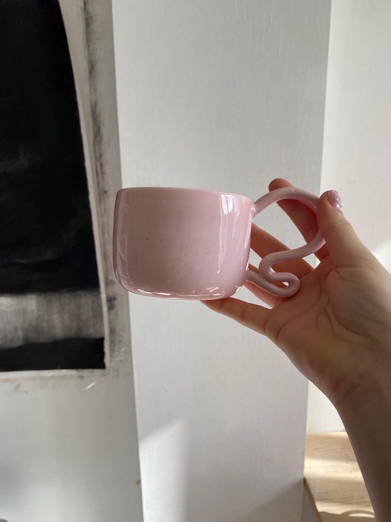 K&ouml;nnte beinhalten: Eine rosa Keramiktasse mit quadratischem Korpus und einem einzigartigen, gewirbelten Henkel. Die Tasse hat eine gesprenkelte Textur und ein gl&auml;nzendes Finish. Die Tasse wird in einer Hand gehalten.