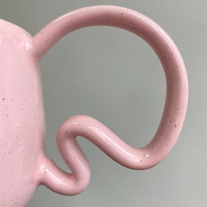 K&ouml;nnte beinhalten: Nahaufnahme einer rosa Keramiktasse mit einem einzigartigen, geschwungenen Henkel. Der Henkel hat ein skurriles, flie&szlig;endes Design. Die Oberfl&auml;che der Tasse ist mit kleinen, dunklen Flecken &uuml;bers&auml;t, die der Gestaltung Textur verleihen.