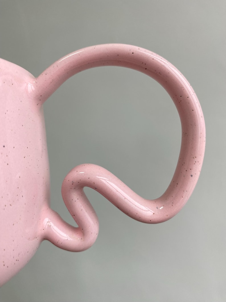 K&ouml;nnte beinhalten: Nahaufnahme einer rosa Keramiktasse mit einem einzigartigen, geschwungenen Henkel. Der Henkel hat ein skurriles, flie&szlig;endes Design. Die Oberfl&auml;che der Tasse ist mit kleinen, dunklen Flecken &uuml;bers&auml;t, die der Gestaltung Textur verleihen.