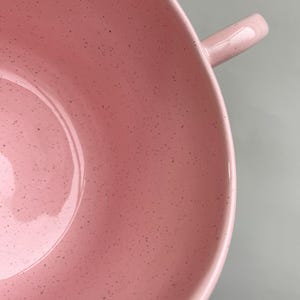 K&ouml;nnte beinhalten: Nahaufnahme einer rosa Keramiksch&uuml;ssel mit Henkel. Die Sch&uuml;ssel hat eine gesprenkelte Textur und eine gl&auml;nzende Oberfl&auml;che. Der Henkel ist eine kleine, gebogene Verl&auml;ngerung des Sch&uuml;sselrandes.
