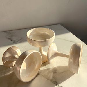 May include: Three ceramic coupe glasses with a glossy, iridescent cream and beige marbled finish. Each glass features a wide, shallow bowl set atop a slender stem and a circular base, showcasing a smooth, reflective surface with subtle color variations.