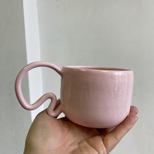K&ouml;nnte beinhalten: Eine rosa Keramik-Tasse mit einem einzigartigen, geschwungenen Henkel. Die Tasse hat eine gesprenkelte Textur und einen abgerundeten K&ouml;rper. Der Griff hat eine wellenf&ouml;rmige, fast schlangenartige Form. Die Tasse wird in einer Hand gehalten, um ihre Gr&ouml;&szlig;e und ihr Design zu zeigen.
