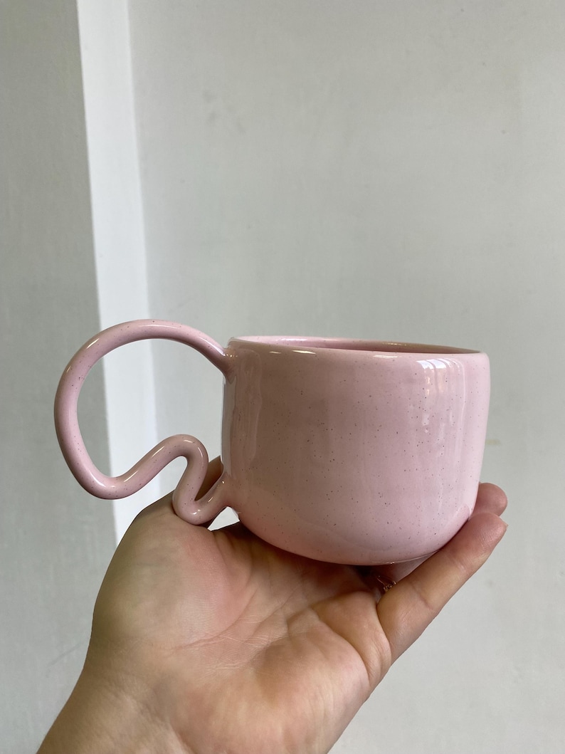 K&ouml;nnte beinhalten: Eine rosa Keramik-Tasse mit einem einzigartigen, geschwungenen Henkel. Die Tasse hat eine gesprenkelte Textur und einen abgerundeten K&ouml;rper. Der Griff hat eine wellenf&ouml;rmige, fast schlangenartige Form. Die Tasse wird in einer Hand gehalten, um ihre Gr&ouml;&szlig;e und ihr Design zu zeigen.
