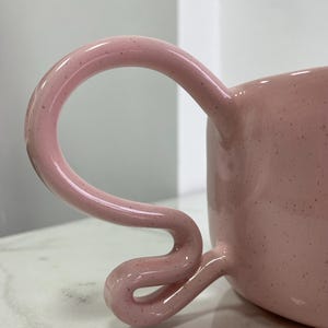 K&ouml;nnte beinhalten: Eine rosa Keramiktasse mit einem einzigartigen, geschwungenen Henkel. Die Tasse hat eine gesprenkelte Textur und ein gl&auml;nzendes Finish. Der Griff hat ein unverwechselbares, schlaufenf&ouml;rmiges Design.