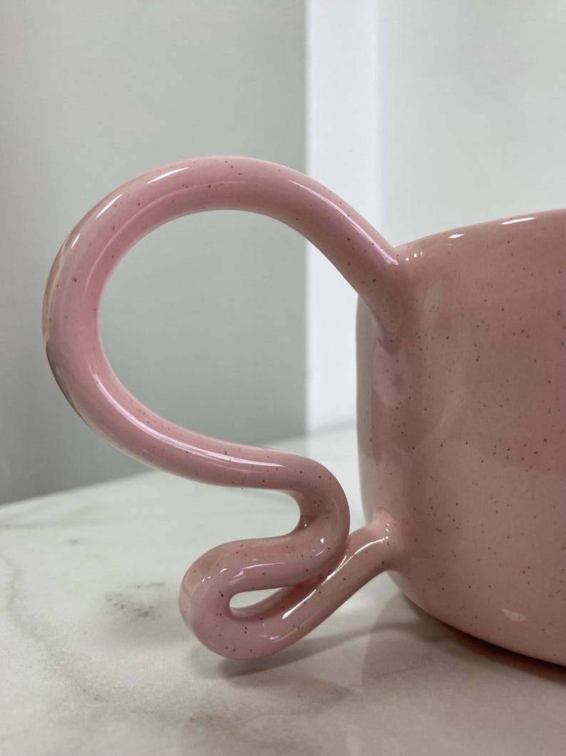 K&ouml;nnte beinhalten: Eine rosa Keramiktasse mit einem einzigartigen, geschwungenen Henkel. Die Tasse hat eine gesprenkelte Textur und ein gl&auml;nzendes Finish. Der Griff hat ein unverwechselbares, schlaufenf&ouml;rmiges Design.