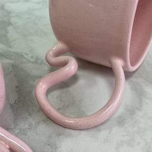 K&ouml;nnte beinhalten: Eine rosa Keramiktasse mit einem einzigartigen, geschwungenen Henkel und gesprenkelter Oberfl&auml;che. Die Tasse liegt auf der Seite und zeigt das markante Griffdesign. Die Gesamt&auml;sthetik ist modern und k&uuml;nstlerisch.