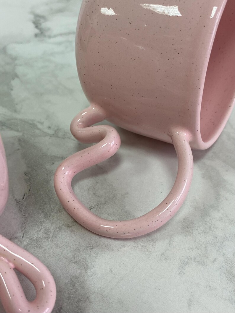 K&ouml;nnte beinhalten: Eine rosa Keramiktasse mit einem einzigartigen, geschwungenen Henkel und gesprenkelter Oberfl&auml;che. Die Tasse liegt auf der Seite und zeigt das markante Griffdesign. Die Gesamt&auml;sthetik ist modern und k&uuml;nstlerisch.