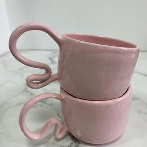 K&ouml;nnte beinhalten: Zwei gestapelte rosa Keramikbecher mit einzigartigen, geschwungenen Griffen. Die Becher haben eine gesprenkelte Textur und eine glatte, gl&auml;nzende Oberfl&auml;che. Die Griffe sind ein markantes Designelement, das der einfachen, abgerundeten Form der Becher einen Hauch von k&uuml;nstlerischem Flair verleiht.