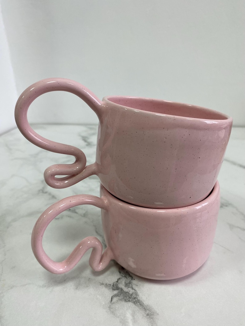 K&ouml;nnte beinhalten: Zwei gestapelte rosa Keramikbecher mit einzigartigen, geschwungenen Griffen. Die Becher haben eine gesprenkelte Textur und eine glatte, gl&auml;nzende Oberfl&auml;che. Die Griffe sind ein markantes Designelement, das der einfachen, abgerundeten Form der Becher einen Hauch von k&uuml;nstlerischem Flair verleiht.