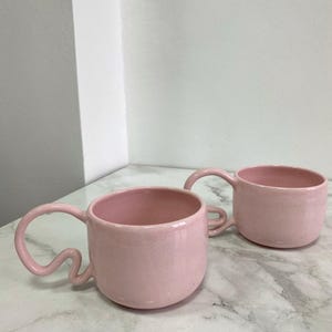 K&ouml;nnte beinhalten: Zwei rosa Keramikbecher mit einzigartigen, geschwungenen Griffen. Die Becher haben eine gesprenkelte Textur und ein gl&auml;nzendes Finish. Die Becher stehen auf einer Marmoroberfl&auml;che. Die Becher sind hellrosa.