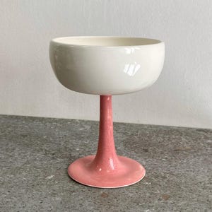 Może przedstawiać: Biała ceramiczna miska z różową nóżką i podstawą. Miska jest okrągła i ma gładkie, błyszczące wykończenie. Nóżka jest wysoka i smukła, a podstawa jest szeroka i płaska.