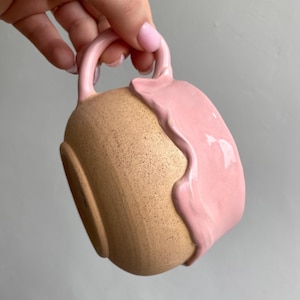 Pode incluir: Uma caneca de cerâmica com uma base de argila bege salpicada e um esmalte rosa liso e brilhante que escorre da borda. A caneca tem uma alça curva e um corpo arredondado, mostrando um contraste entre a argila texturizada e o acabamento.