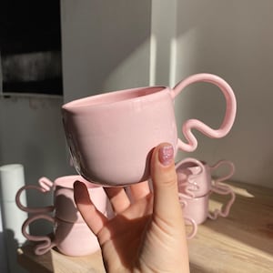 K&ouml;nnte beinhalten: Mehrere rosa Keramikbecher mit einzigartigen, wellenf&ouml;rmigen Griffen. Die Becher sind in einem weichen, pastellrosa gehalten und scheinen handgefertigt zu sein. Die Becher stehen auf einer Holzoberfl&auml;che.