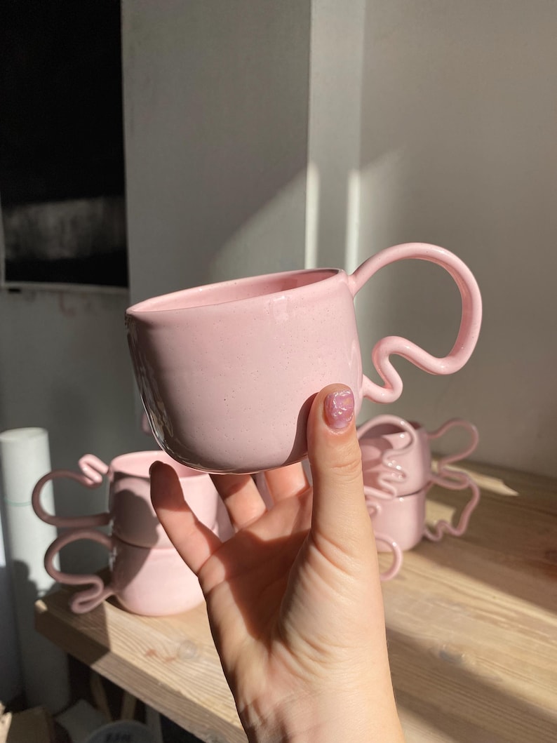 K&ouml;nnte beinhalten: Mehrere rosa Keramikbecher mit einzigartigen, wellenf&ouml;rmigen Griffen. Die Becher sind in einem weichen, pastellrosa gehalten und scheinen handgefertigt zu sein. Die Becher stehen auf einer Holzoberfl&auml;che.