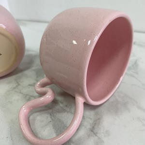 K&ouml;nnte beinhalten: Eine rosa Keramik-Tasse mit einem einzigartigen, geschwungenen Henkel. Die Tasse hat eine gesprenkelte Oberfl&auml;che und ein glattes, gl&auml;nzendes Inneres. Der Henkel ist in einer skurrilen, fast serpentinenartigen Form gebogen. Die Tasse ist f&uuml;r Getr&auml;nke konzipiert.