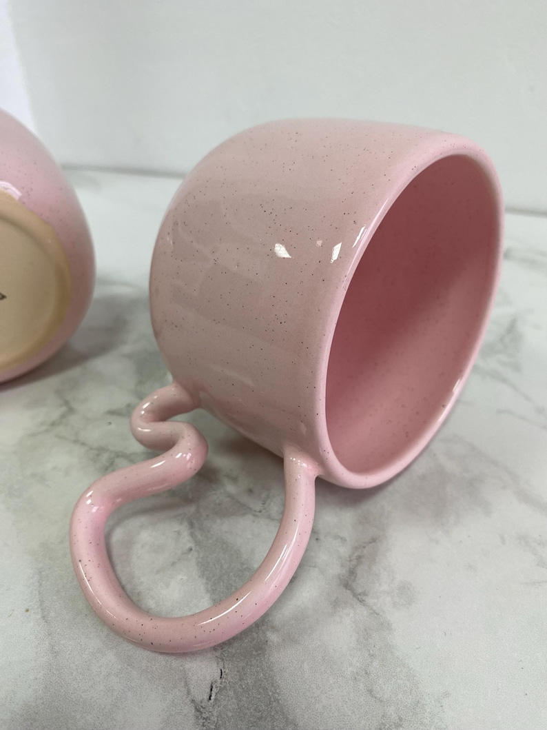 K&ouml;nnte beinhalten: Eine rosa Keramik-Tasse mit einem einzigartigen, geschwungenen Henkel. Die Tasse hat eine gesprenkelte Oberfl&auml;che und ein glattes, gl&auml;nzendes Inneres. Der Henkel ist in einer skurrilen, fast serpentinenartigen Form gebogen. Die Tasse ist f&uuml;r Getr&auml;nke konzipiert.